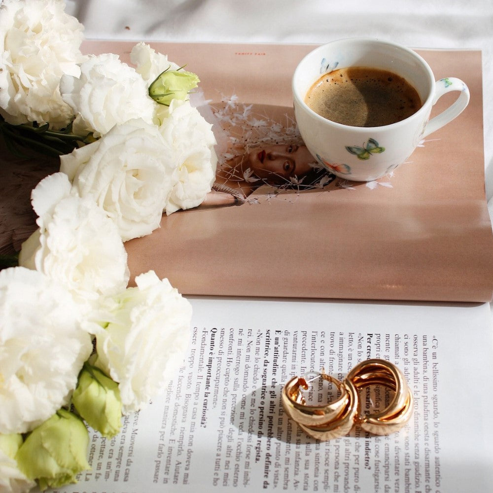 Gold jewelry sits on a table next to some flowers and coffee