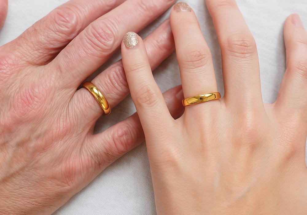 Two hands placed side by side, each wearing a gold wedding band. The hand on the left is an older hand, and the hand on the right is a younger hand, both displaying smooth, polished rings